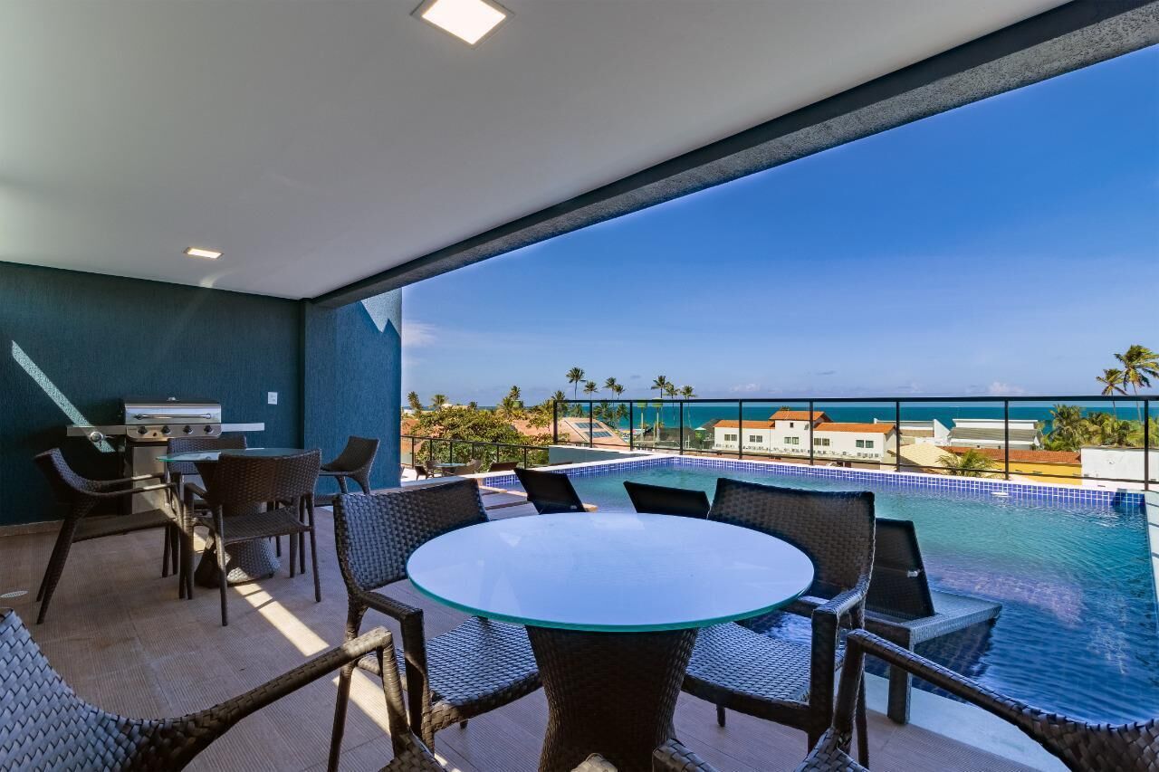 A balcony with a glass table and chairs, a grill, and a view of the ocean and buildings.