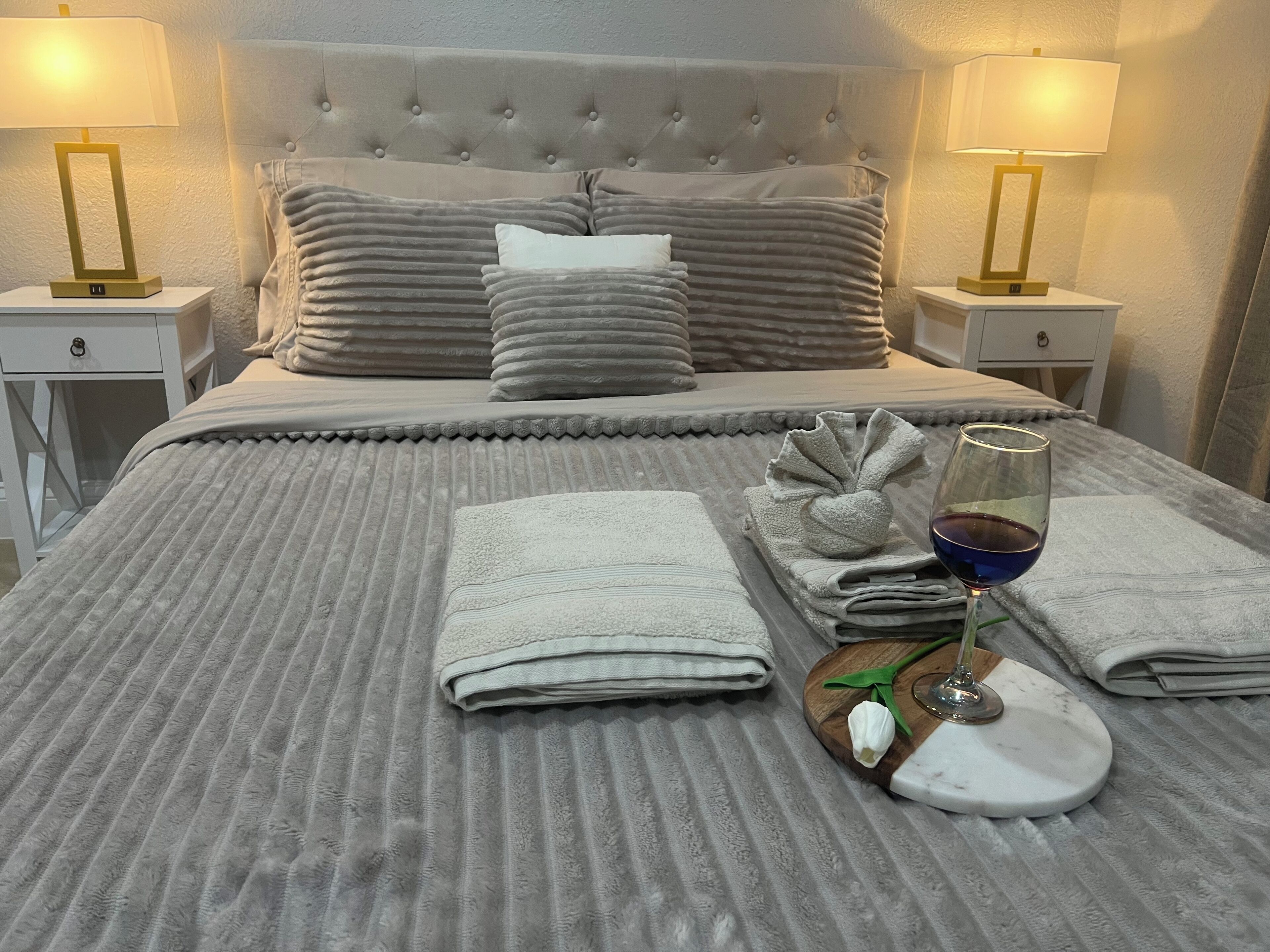A neatly made bed with grey bedding, two bedside tables with lamps, and a glass of red wine on a coaster.