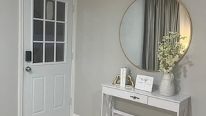 En hall med en vit dörr, en rund spegel med en guldfärgad ram, ett vitt konsolbord med en vas med blommor och en skylt med texten ”Välsignad”.