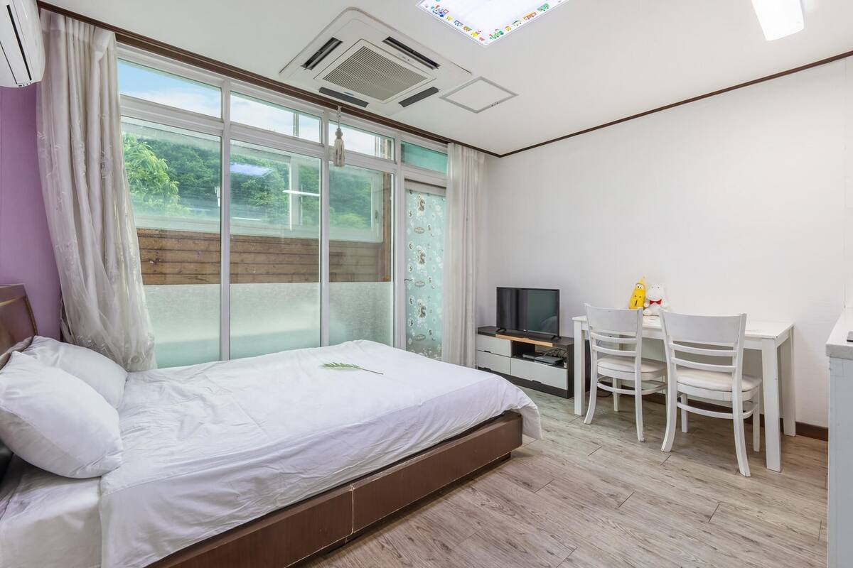 A modern hotel room with a large bed, a desk with a chair, a television, and a small kitchenette.