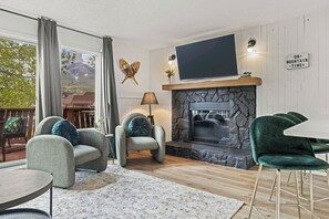 A living room with a stone fireplace, a flat-screen TV, and a view of mountains through large windows.
