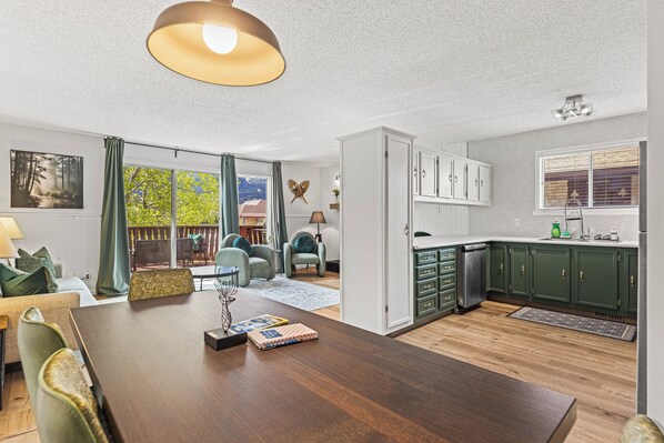 Een ruime woonkamer met een houten eettafel, groene beklede stoelen en uitzicht op een weelderig groen landschap door grote ramen.