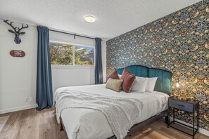 Een slaapkamer met een groot bed, een hoofdeinde, een nachtkastje, een aan de muur bevestigd hertenhoofd en een raam met gordijnen, plus behang met een patroon.