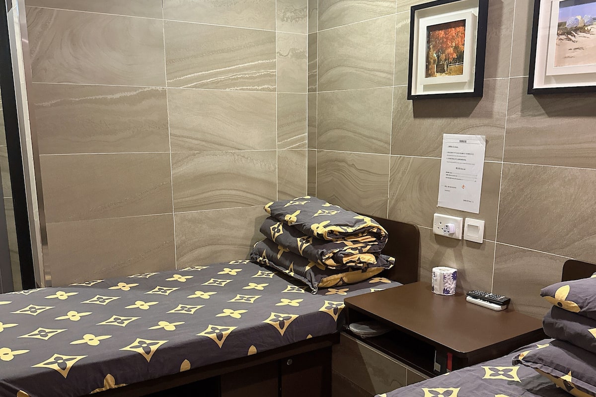 A hotel room with two beds, each with a patterned bedspread, a small desk with a remote control, and a framed picture on the wall.