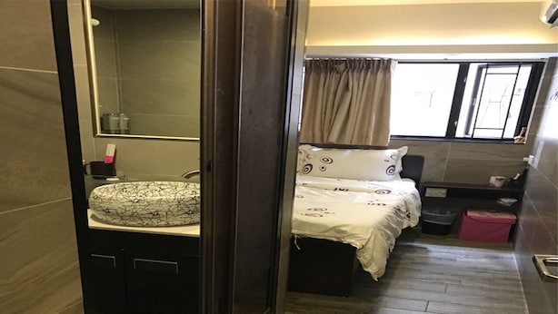 A compact hotel room with a bed, a small bedside table, a mirror, and a window with curtains. - Zixin Hotel (Kowloon)