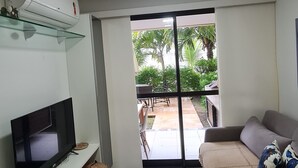 A living room with a flat-screen TV, a glass shelf, a sofa, and a sliding glass door leading to an outdoor patio. - Cupe Beach Living by La Playa (Ipojuca)