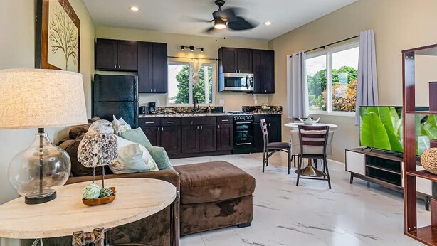 A modern kitchen with dark cabinets, a black refrigerator, and a microwave. There is a dining area with a round table and a brown sofa. - Experience Tropical Tranquility: Luxury Studio Home in Hilo!  (Hilo)