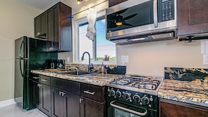 A modern kitchen with dark wood cabinets, stainless steel appliances, a granite countertop, and a window with a view of greenery. - Experience Tropical Tranquility: Luxury Studio Home in Hilo!  (Hilo)