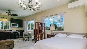 A bedroom with a large bed, a bedside table with a lamp, a window with curtains, and a view of the outside. - Experience Tropical Tranquility: Luxury Studio Home in Hilo!  (Hilo)
