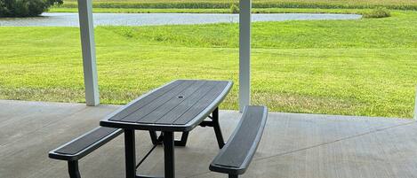 A picnic table with benches under a covered area with large windows overlooking a grassy field and a body of water.