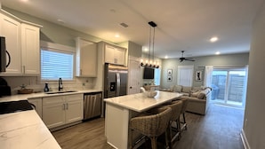 A modern kitchen with white cabinets, stainless steel appliances, a large island, and a dining area with bar stools. - New Condo, Sleeps 6, 3 Bedroom, Beach Access, Community Pool & Private Cabanas. (Corolla)