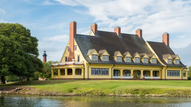 A large, yellow building with multiple chimneys, a pond in front, and a large tree to the left. - New Condo, Sleeps 6, 3 Bedroom, Beach Access, Community Pool & Private Cabanas. (Corolla)