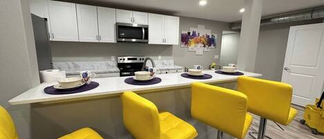 A modern kitchen with white cabinets, a stainless steel microwave, and a bar area with yellow chairs.