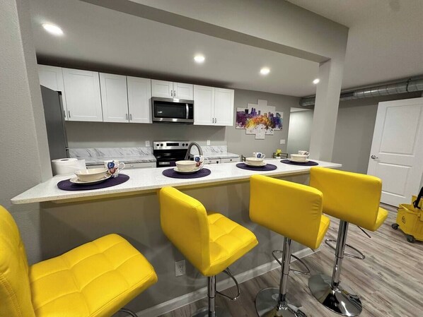 A modern kitchen with white cabinets, a stainless steel microwave, and a bar area with yellow chairs.