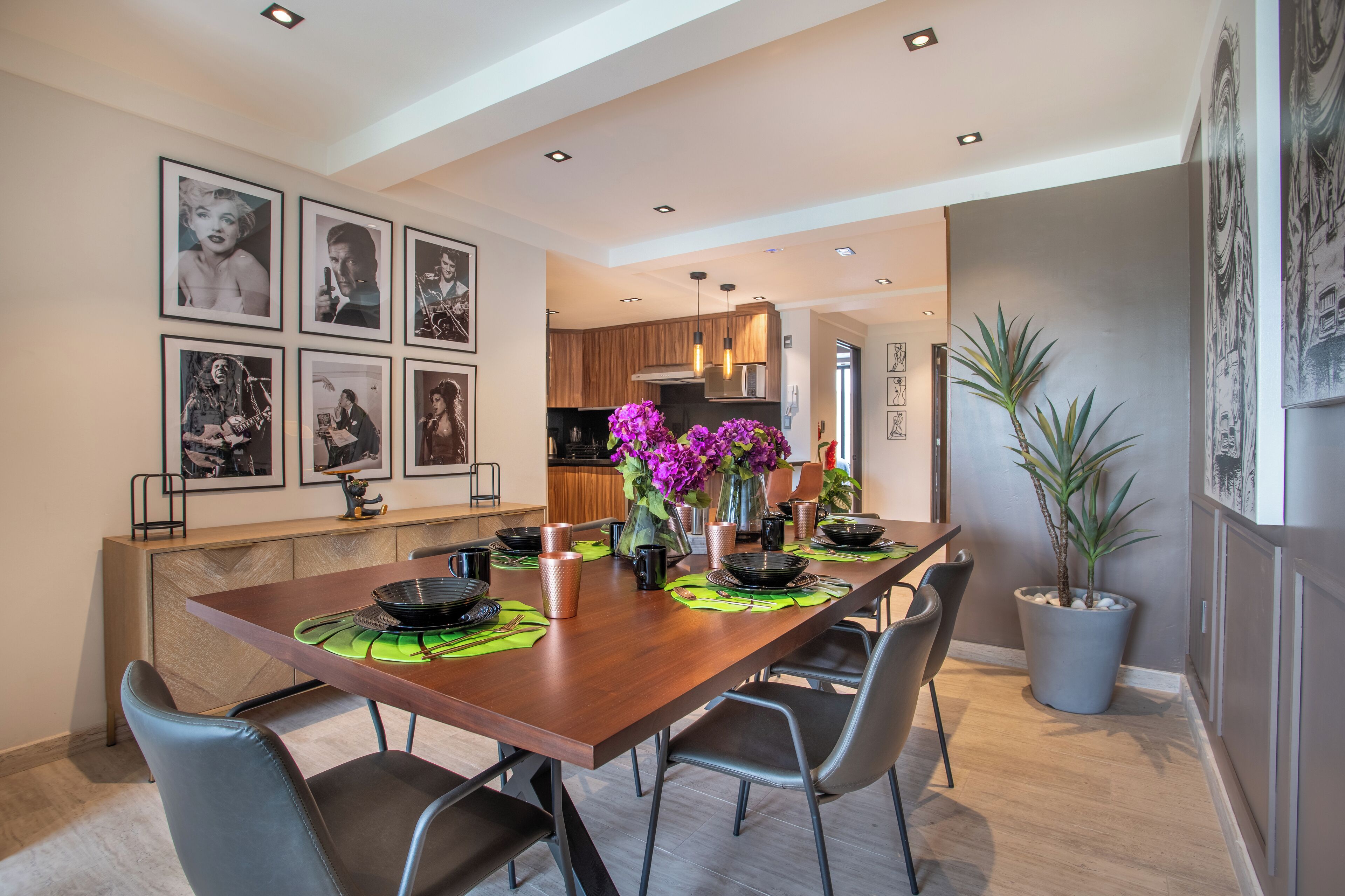 Un comedor moderno con una mesa de madera, individuales negros y verdes, y un jarrón con flores moradas.