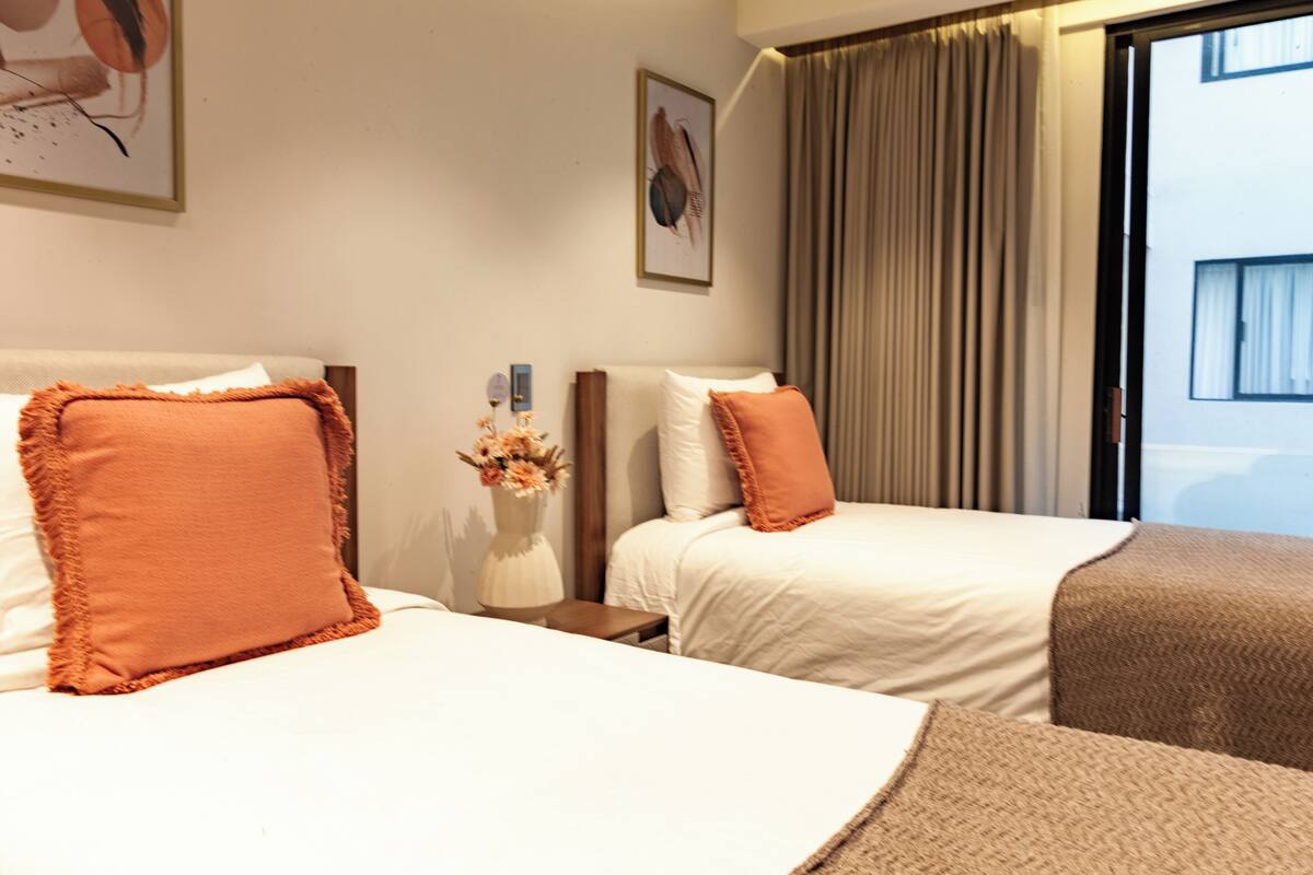 Habitación de hotel con dos camas, una mesita de noche con un jarrón de flores y un ventanal con cortinas.