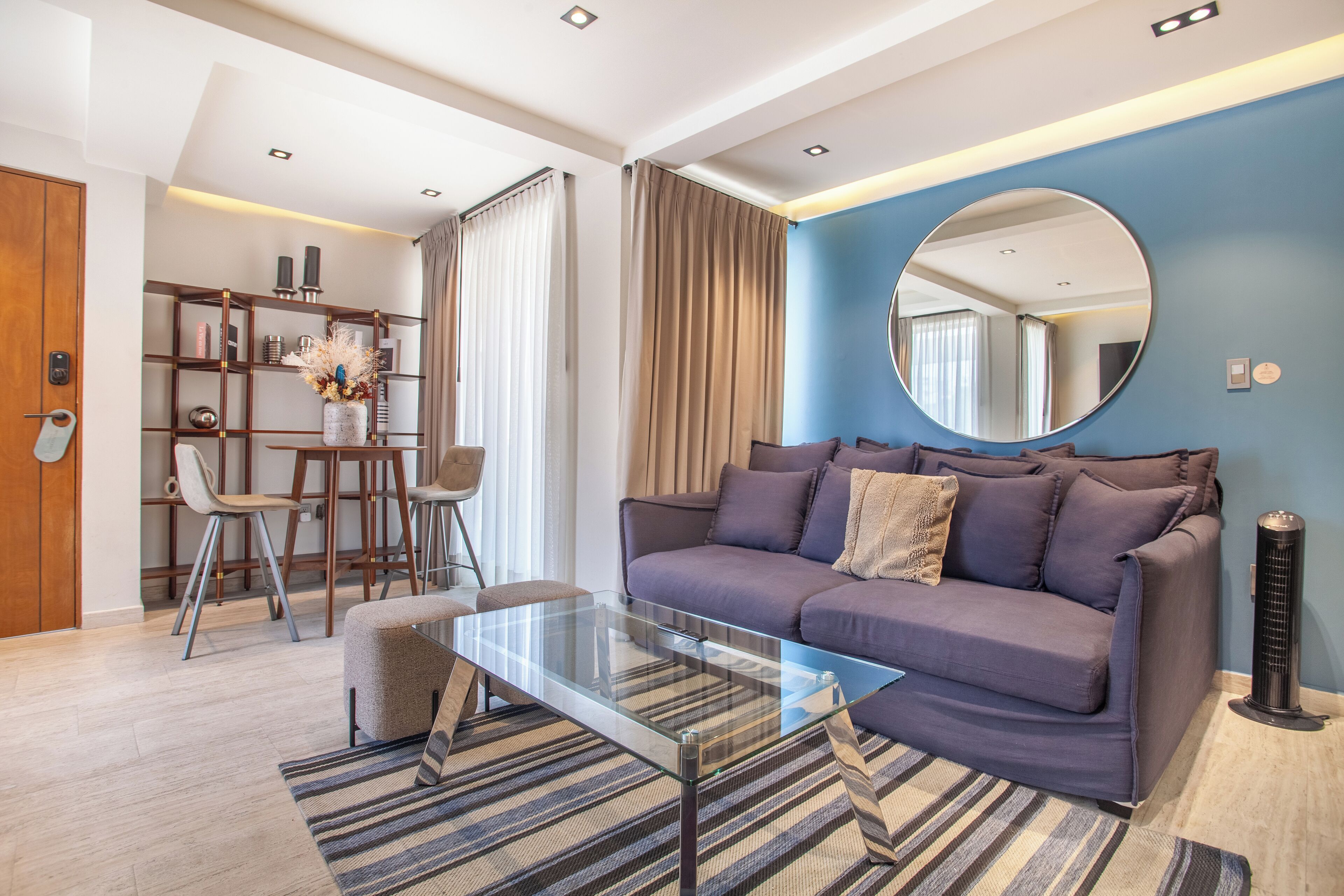 A modern living room with a glass coffee table, a sofa, a round mirror, and a bookshelf.