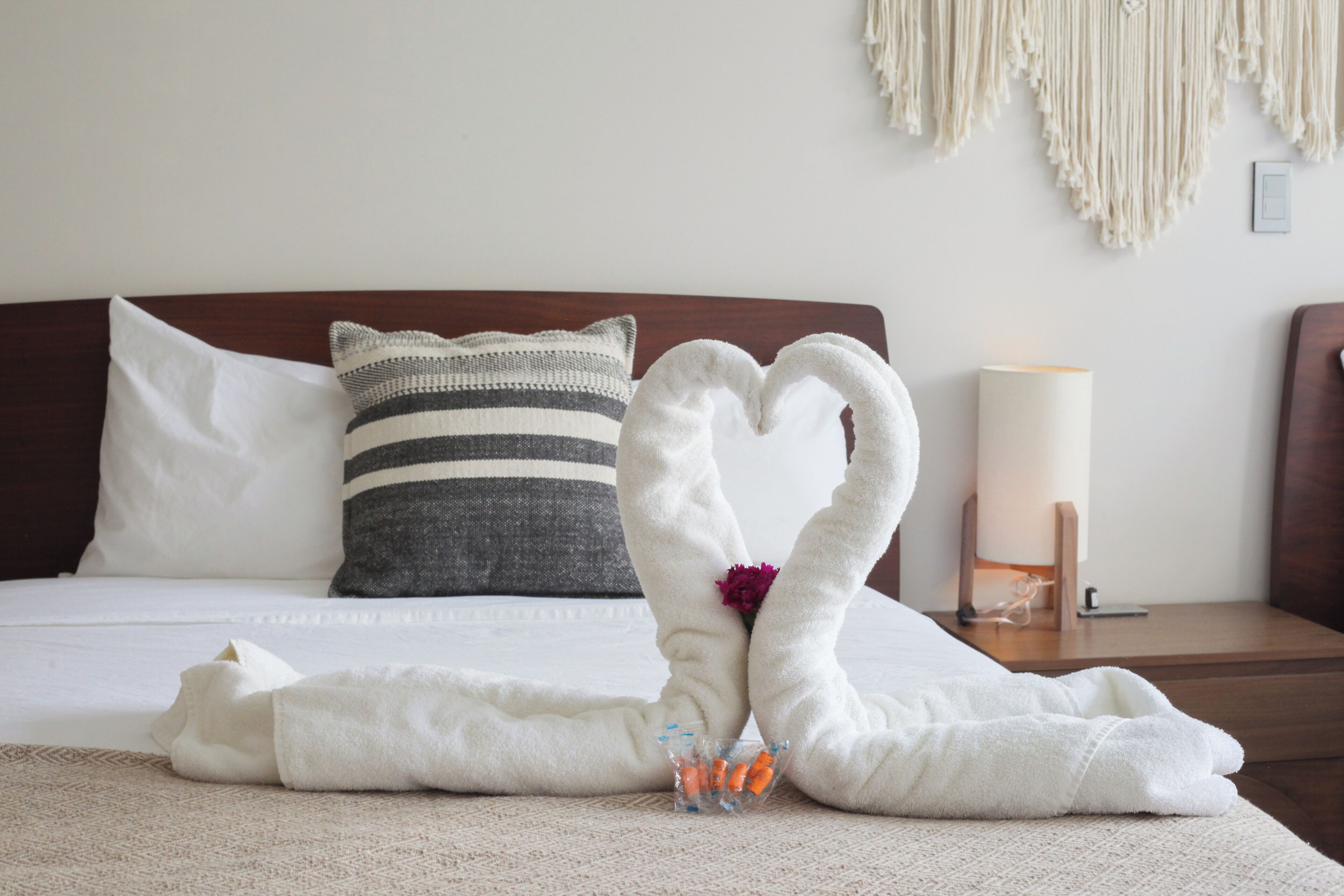 Una cama bien hecha con una toalla dispuesta en forma de corazón, una almohada a rayas y una lámpara de noche.