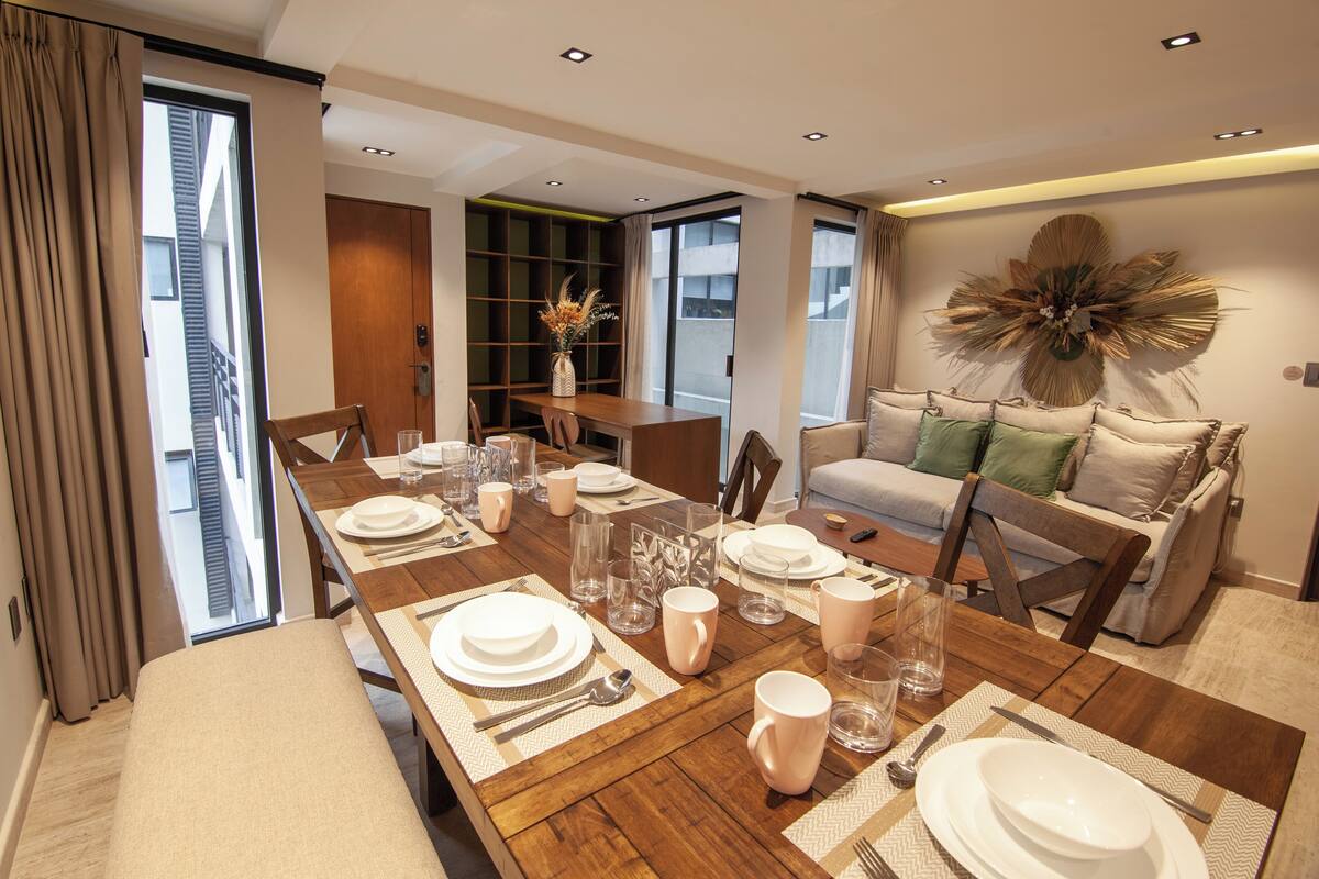 Un comedor moderno con una mesa de madera preparada para cuatro personas, un sofá con cojines y una estantería.
