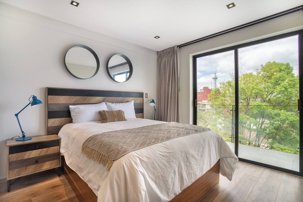 Un dormitorio moderno con una cama grande, dos mesitas de noche, un espejo y vistas a la vegetación a través de amplias ventanas.