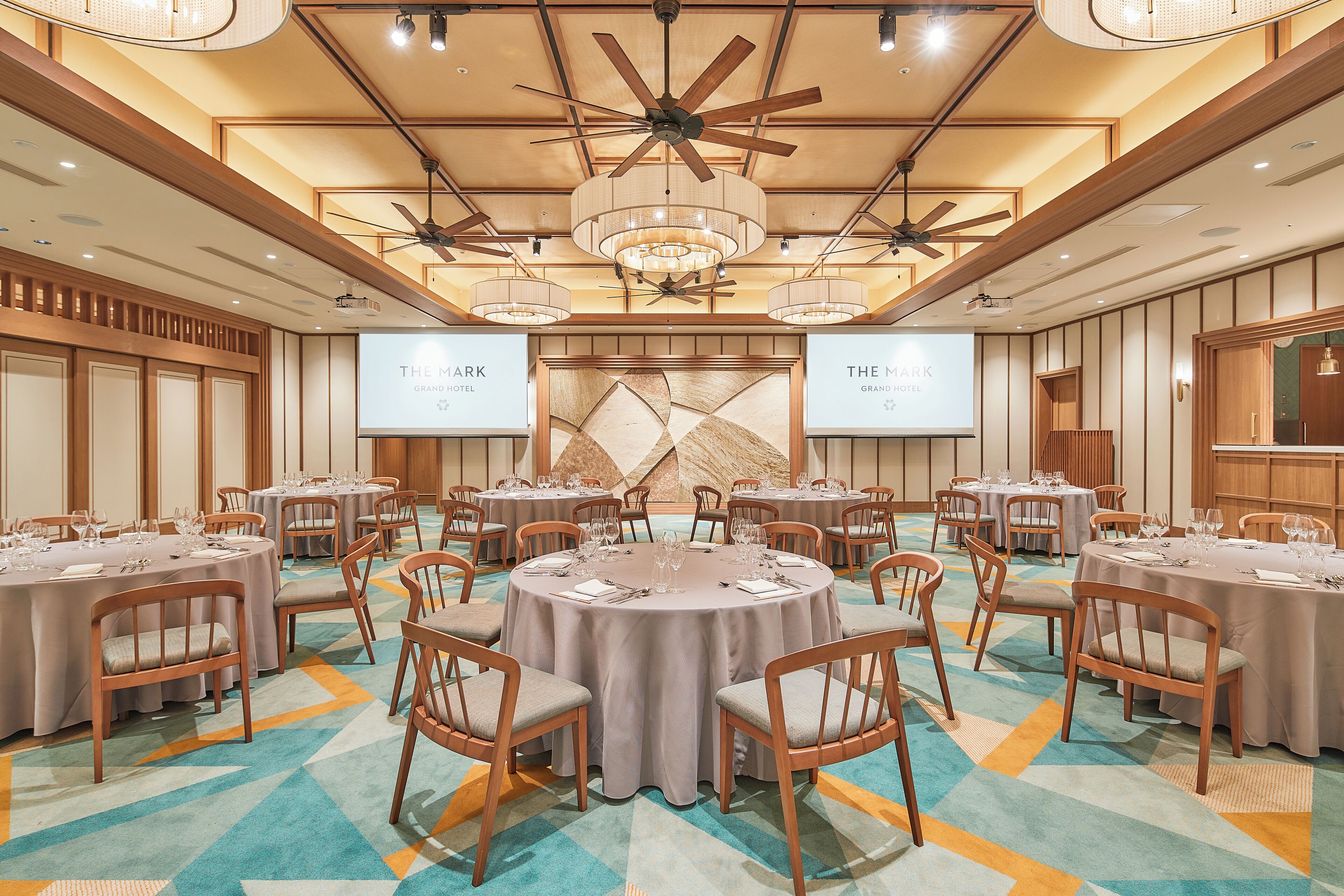 Ein Konferenzraum mit runden Tischen, Stühlen und einem großen Bildschirm, der "THE MARRIOTT GRAND HOTEL" anzeigt.