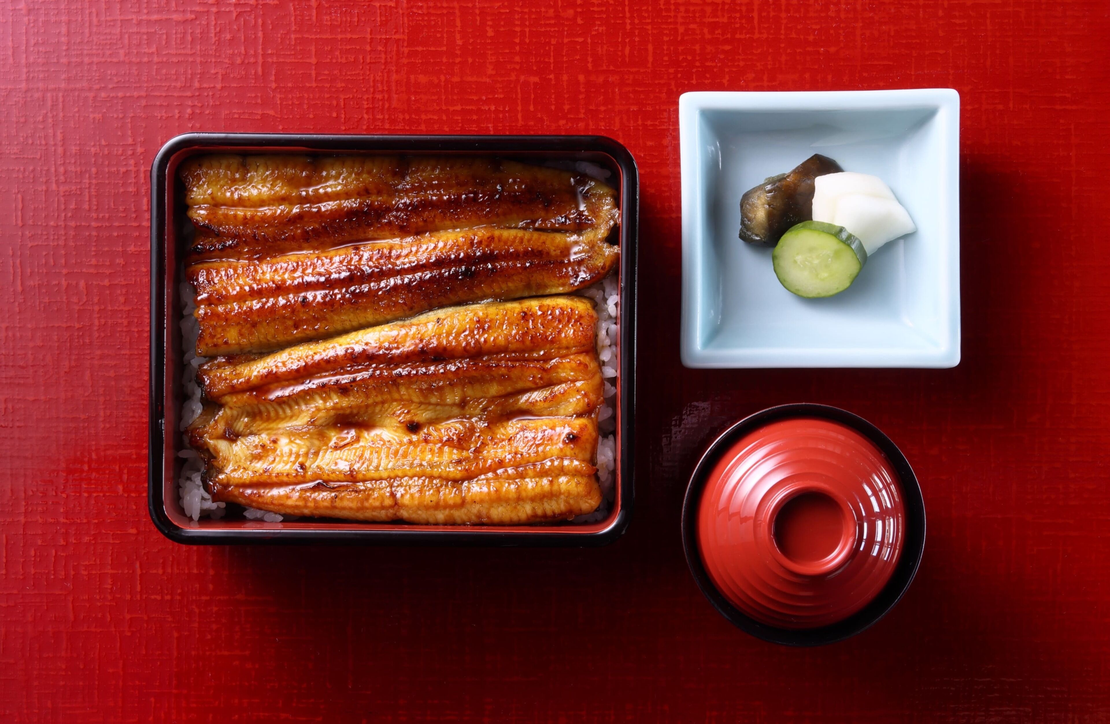 A tray of grilled fish on rice, a small dish with a cucumber and a piece of meat, and a red cup on a red table.