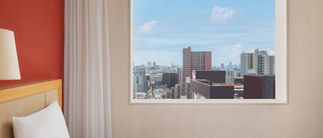 A hotel room with a bed, a window with a cityscape view, a red wall, and a white curtain.