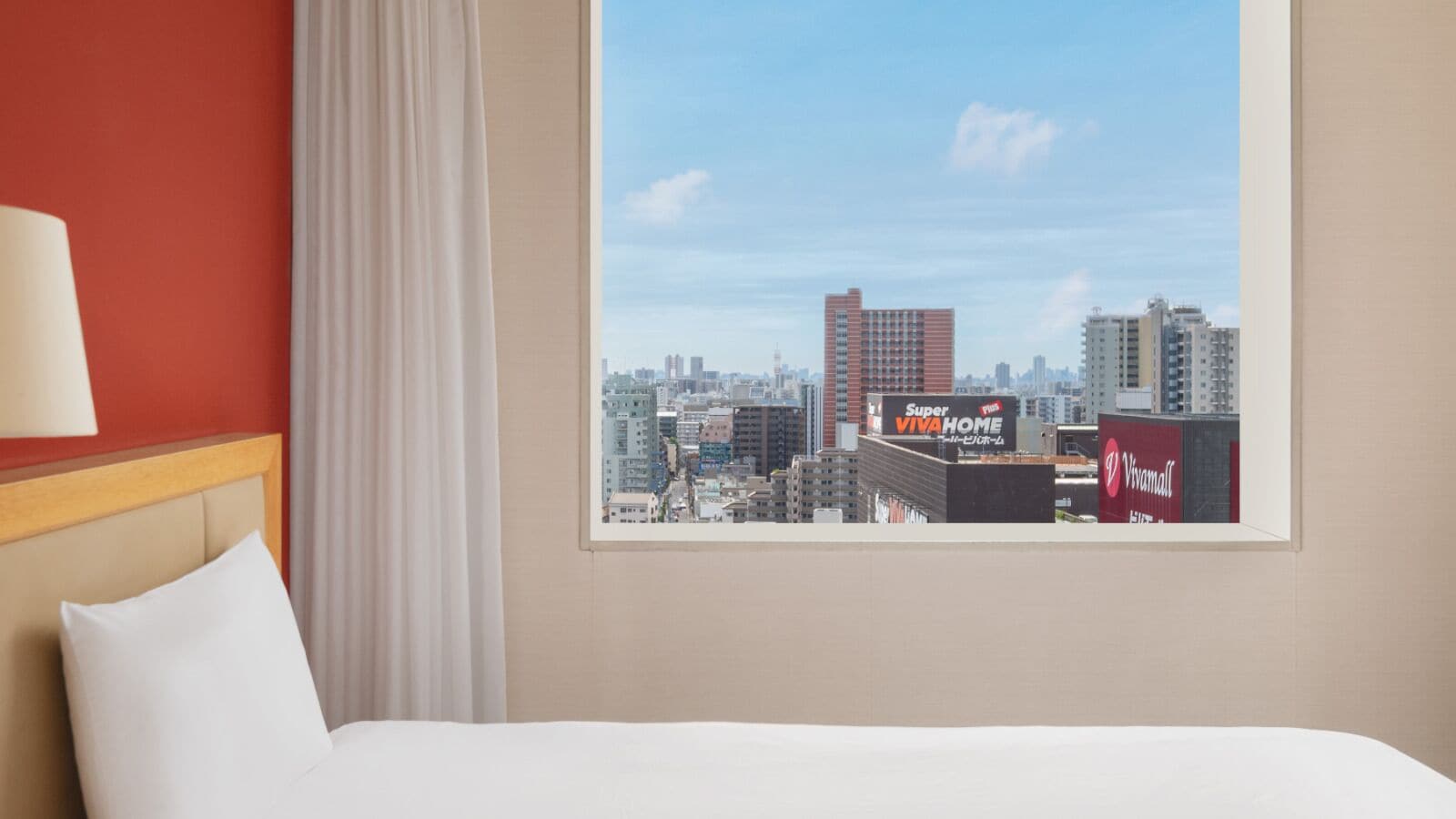 A hotel room with a bed, a window with a cityscape view, a red wall, and a white curtain.
