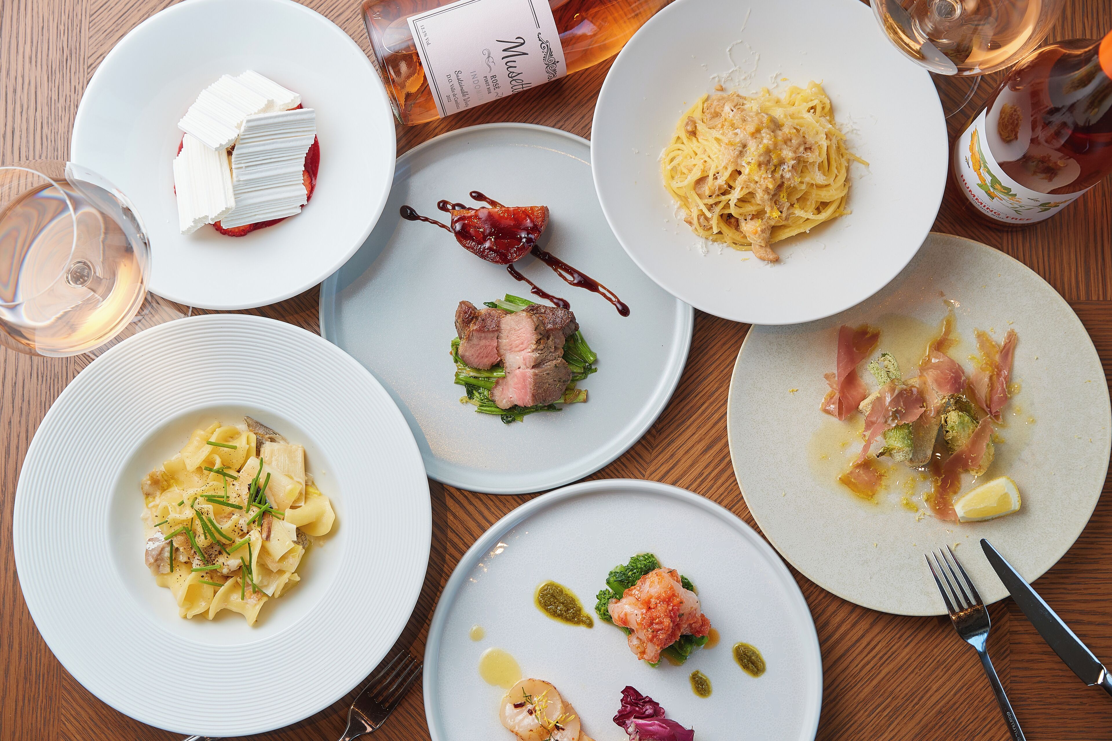 Une variété de plats présentés sur une table en bois, notamment des plats de pâtes, de fruits de mer et de viande, accompagnés de verres à vin et d’une bouteille de vin.