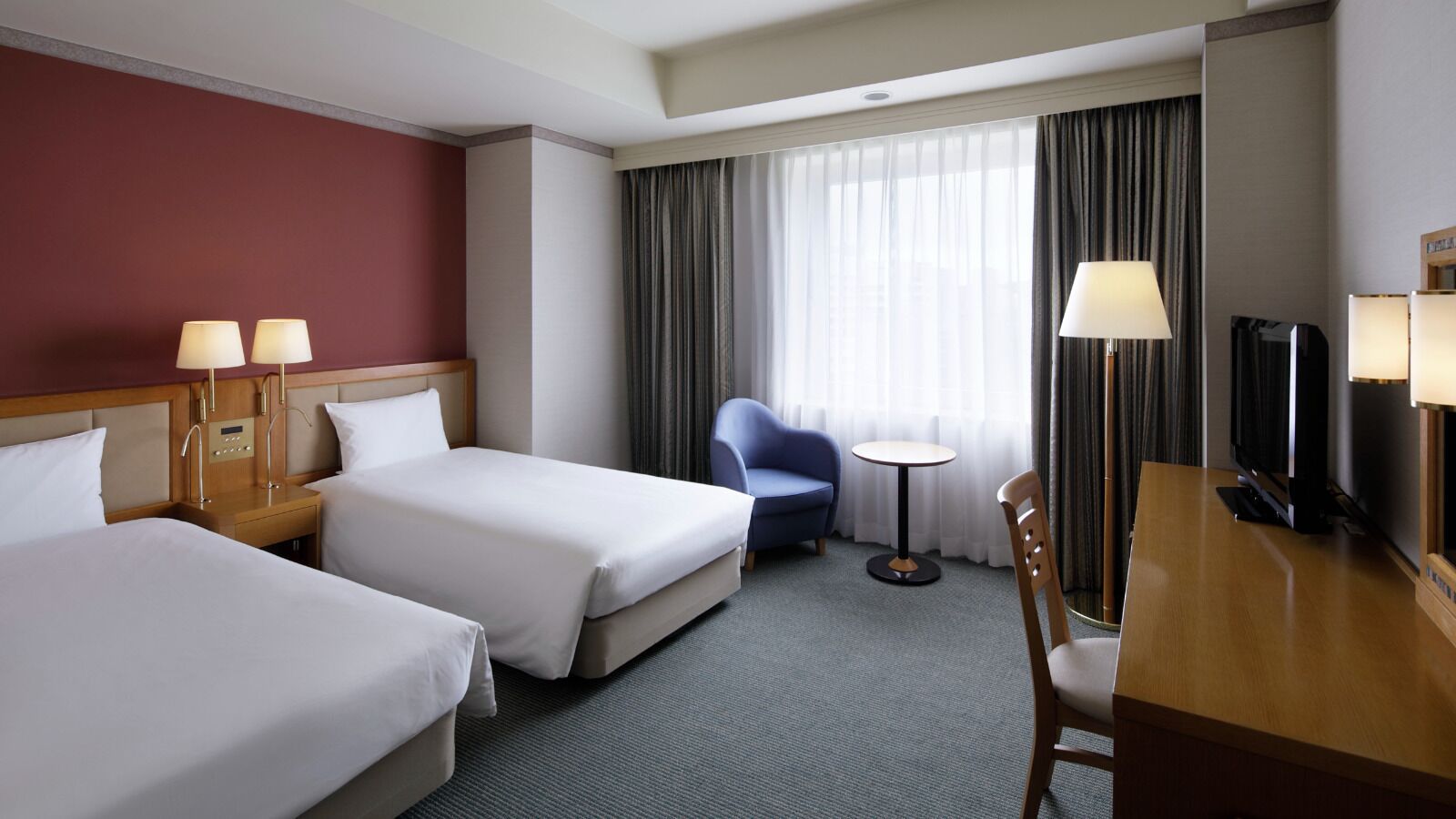 Ein Hotelzimmer mit zwei Betten, einem Schreibtisch, einem Stuhl, einem Fernseher und einem kleinen Tisch mit Lampe.