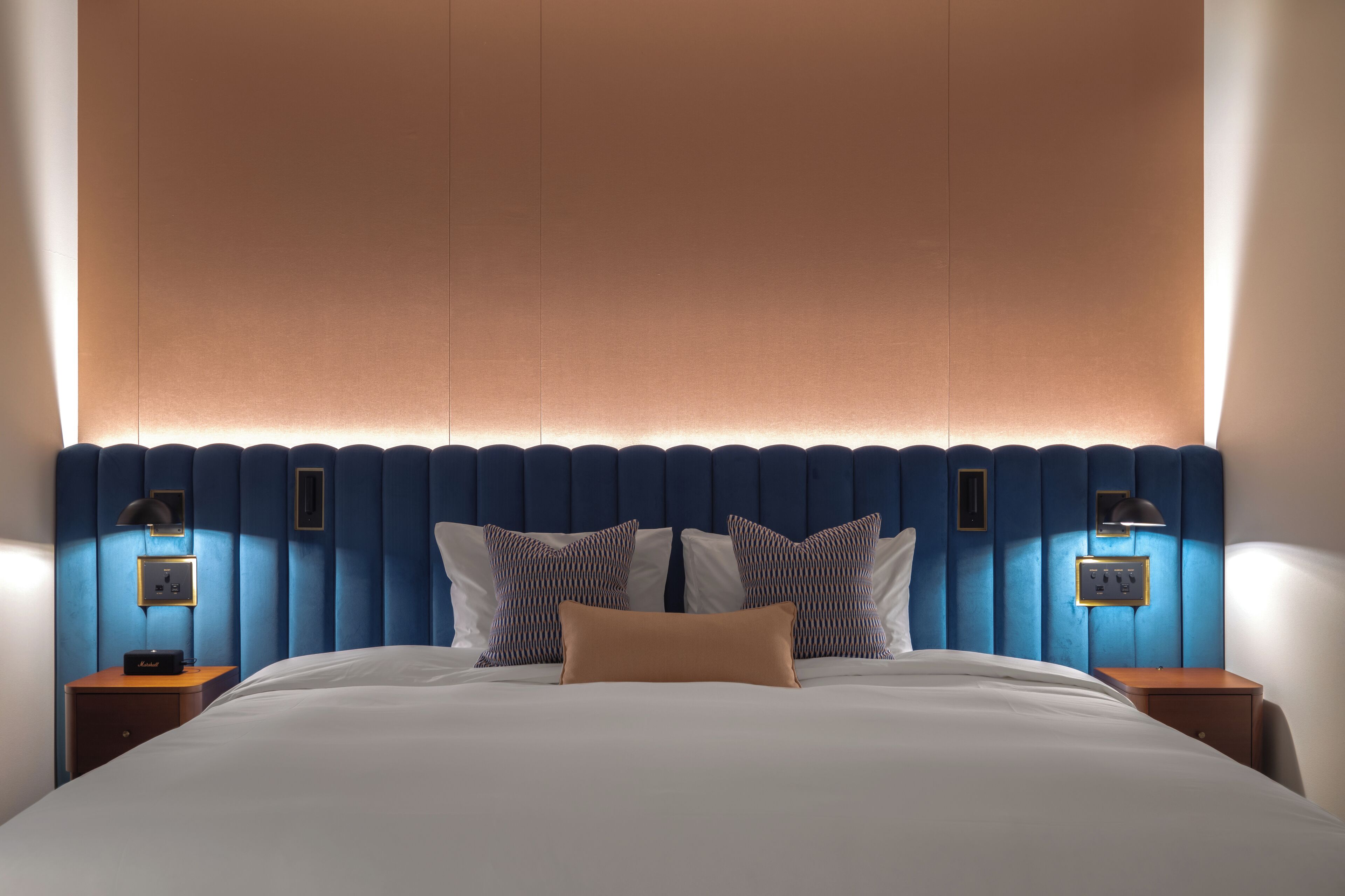A hotel room with a large bed, a blue headboard, two bedside lamps, and a nightstand with a lamp.