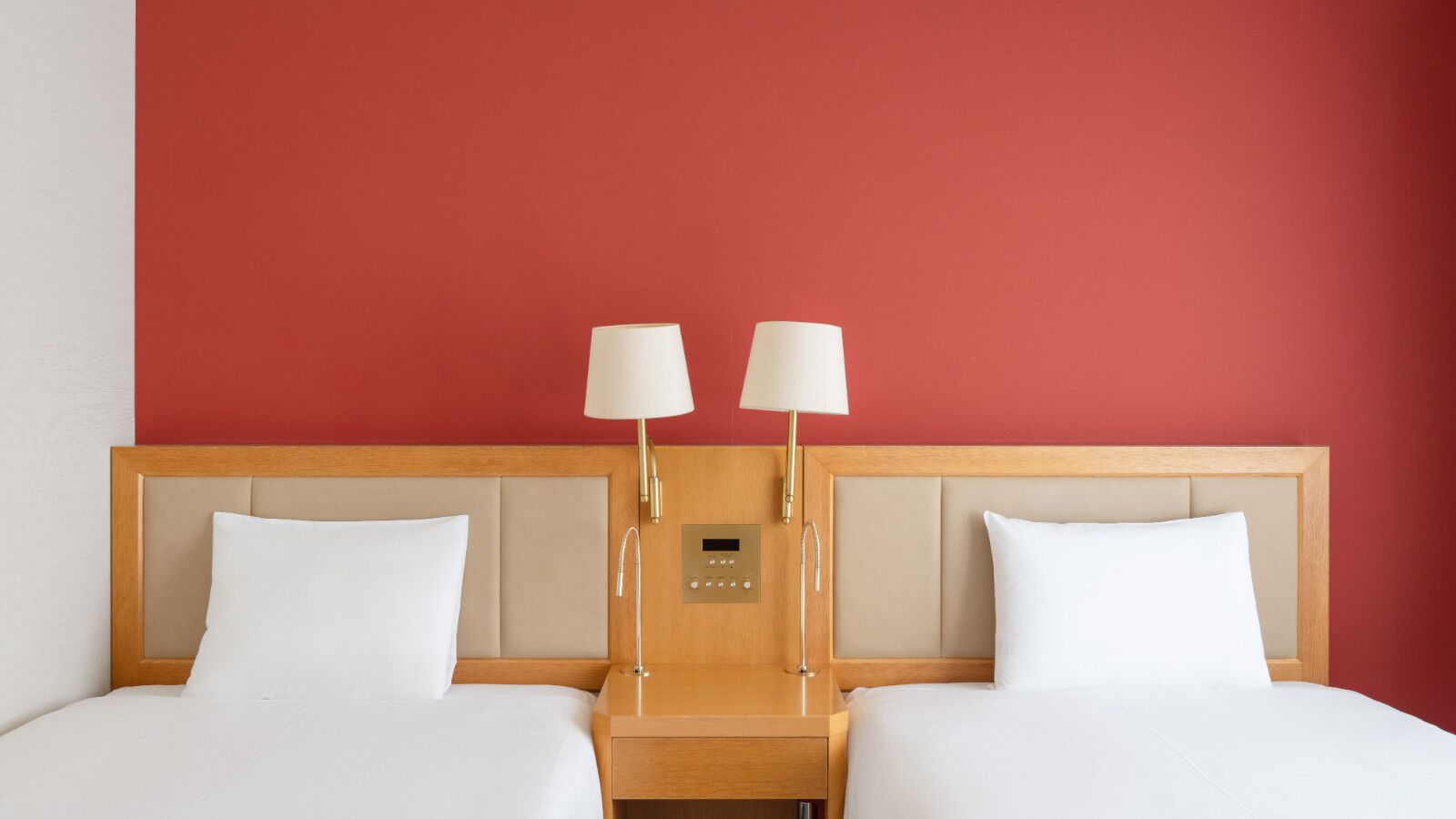 Ein Hotelzimmer mit roter Wand, zwei Betten mit weißen Leinentüchern, einem hölzernen Kopfteil, Nachttischen mit Lampen und einer Bedienkonsole am Kopfteil.