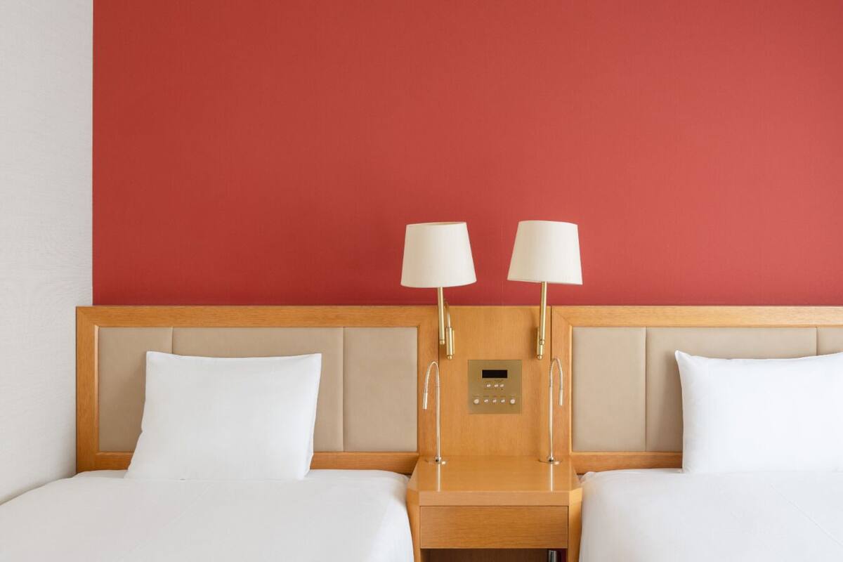 A hotel room with a red wall, two beds with white linens, wooden headboard, bedside tables with lamps, and a control panel on the headboard.