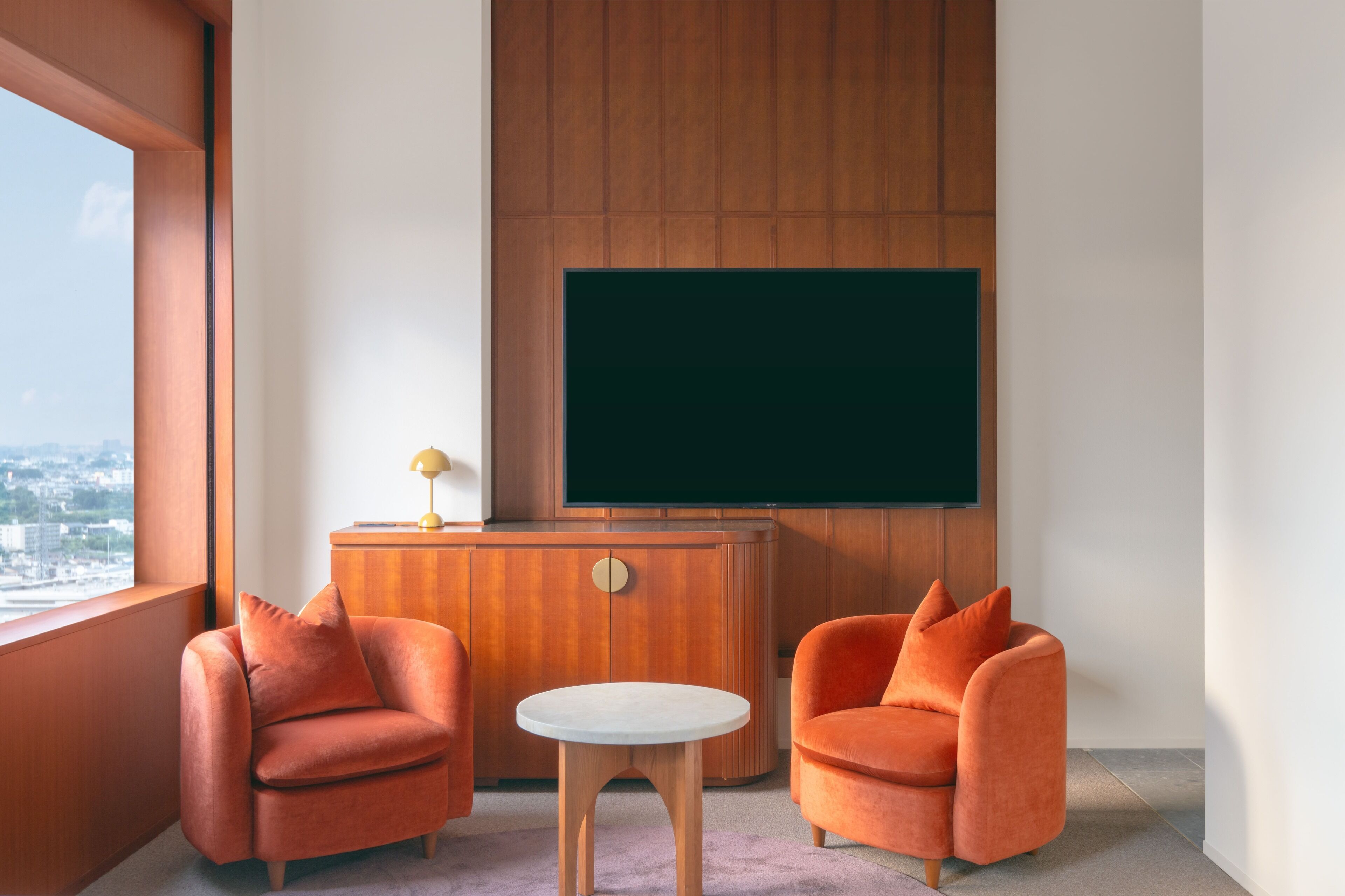 A modern room with two orange armchairs, a small round table, and a wooden cabinet with a television mounted on the wall.