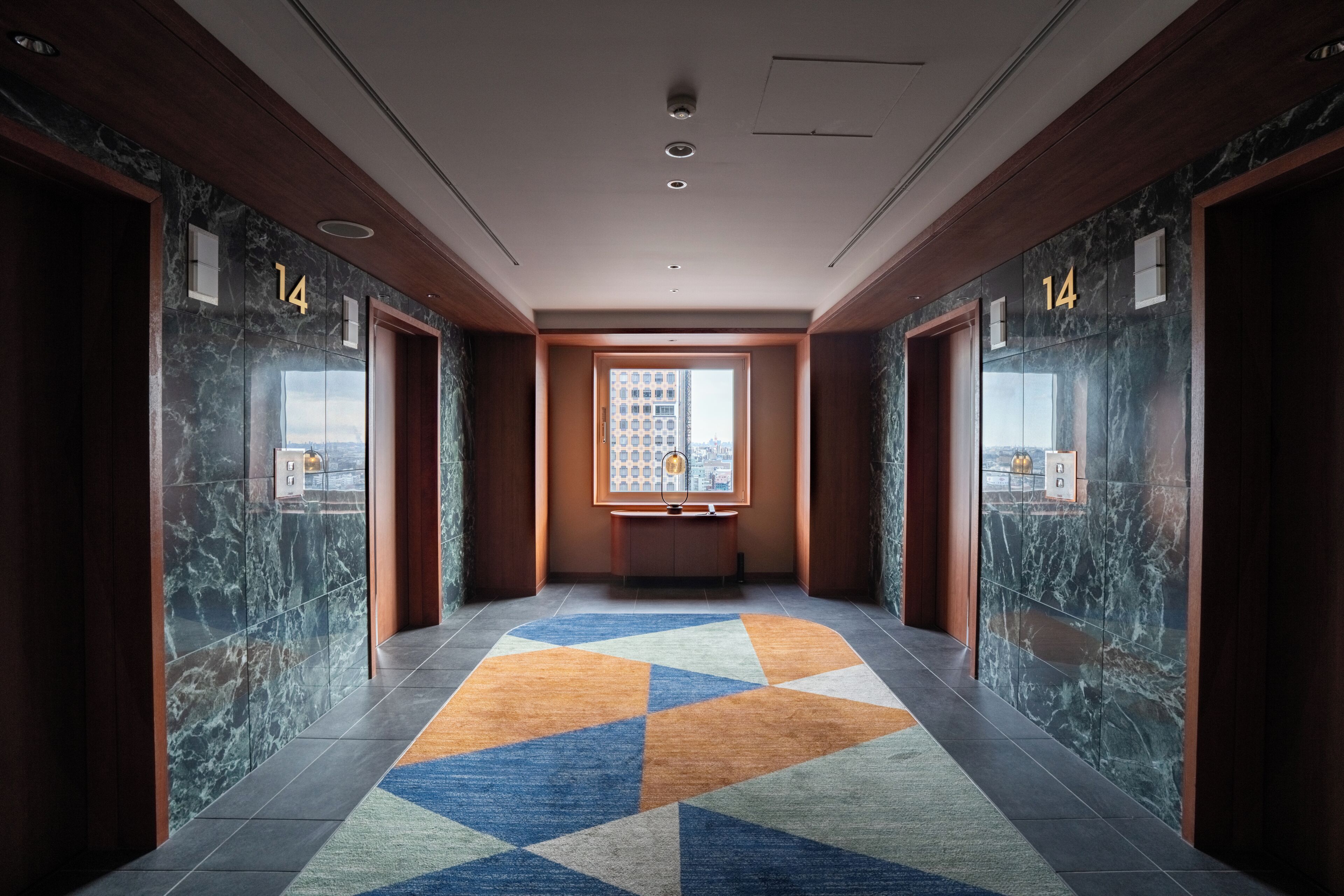 A hallway with marble walls, a geometric patterned carpet, and a view of a cityscape through a window.