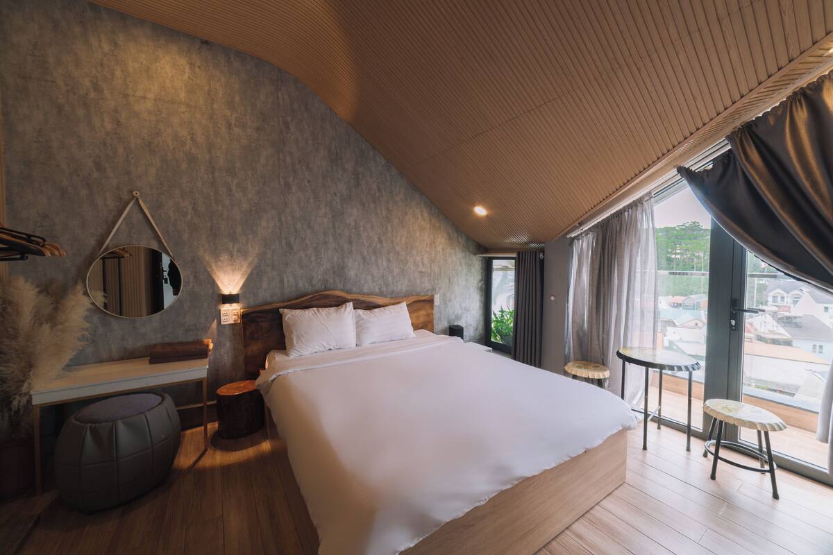 A modern bedroom with a large bed, a wooden headboard, a round mirror, and a view of the outside through large windows.