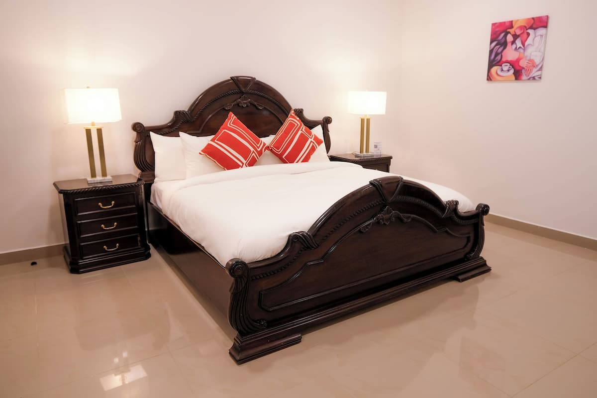 Una cama bien hecha con cabecera de madera, dos mesitas de noche con lámparas y un cuadro colgado en la pared.