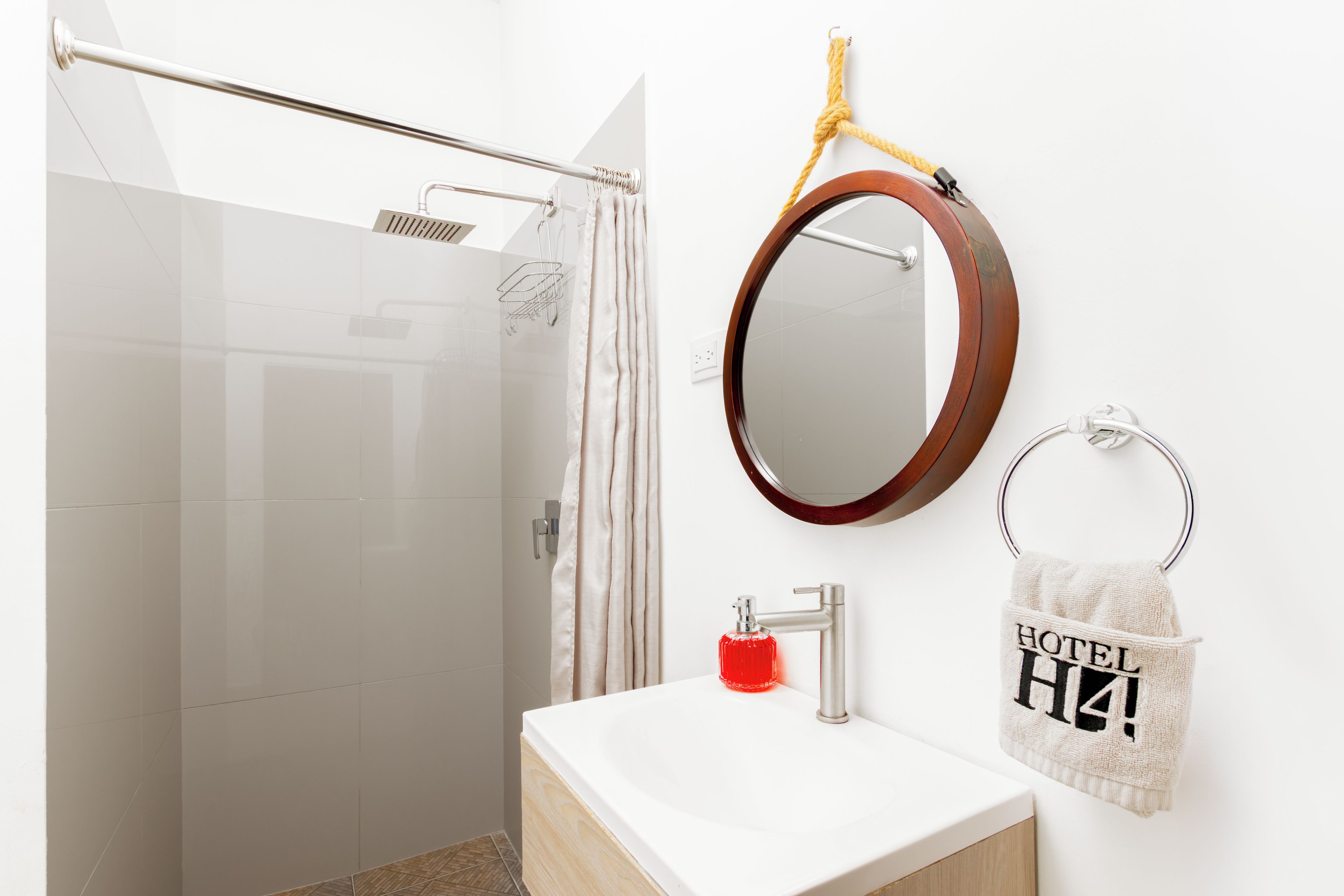 A bathroom with a glass shower enclosure, a white sink with a wooden vanity, a round mirror with a wooden frame, and a towel rack with a towel labeled 'HOTEL'.
