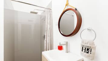 A bathroom with a glass shower enclosure, a white sink with a wooden vanity, a round mirror with a wooden frame, and a towel rack with a towel labeled 'HOTEL'.