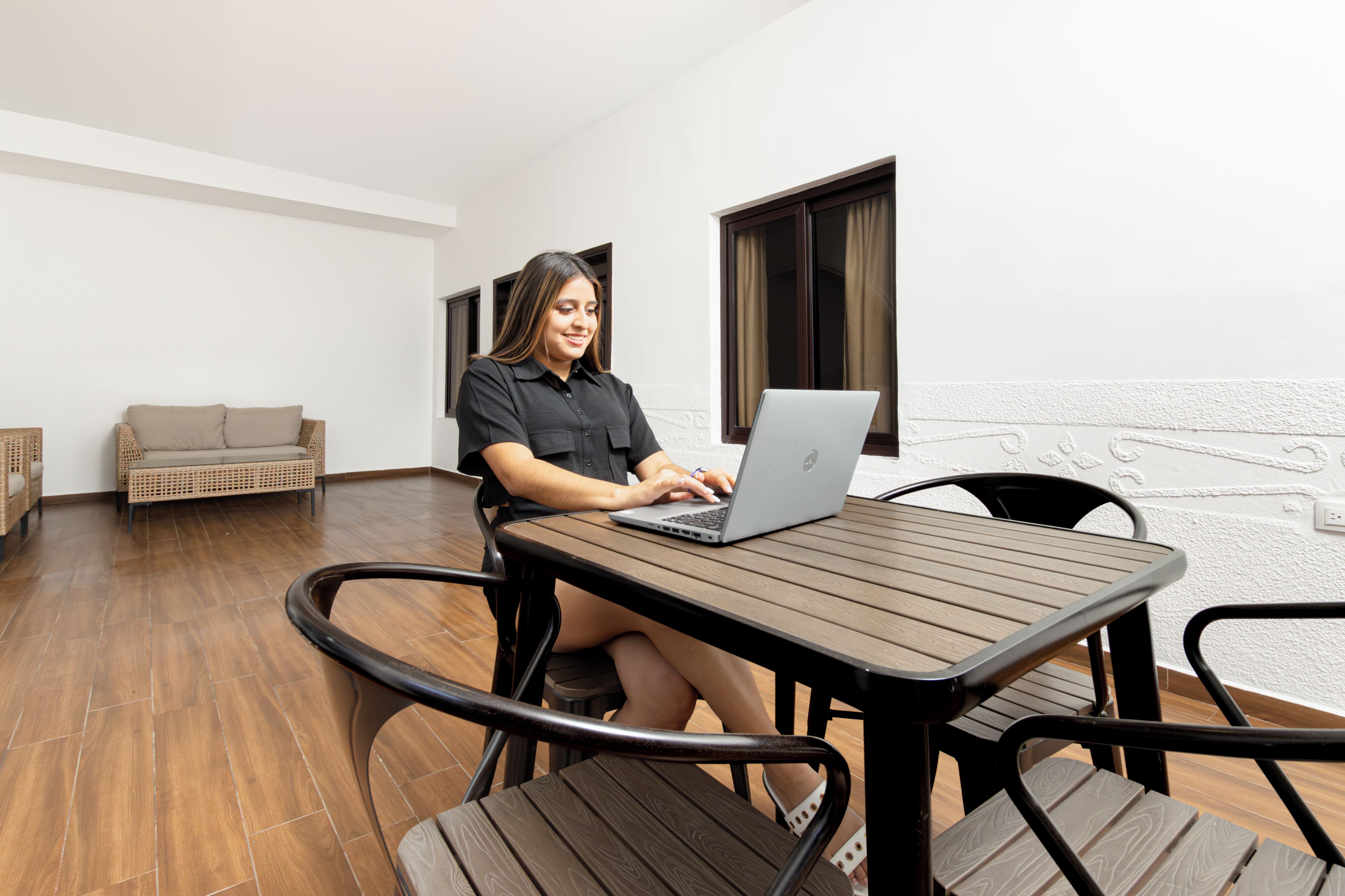 A person seated at a wooden table with a laptop, in a room with a wicker sofa and a patterned wall.