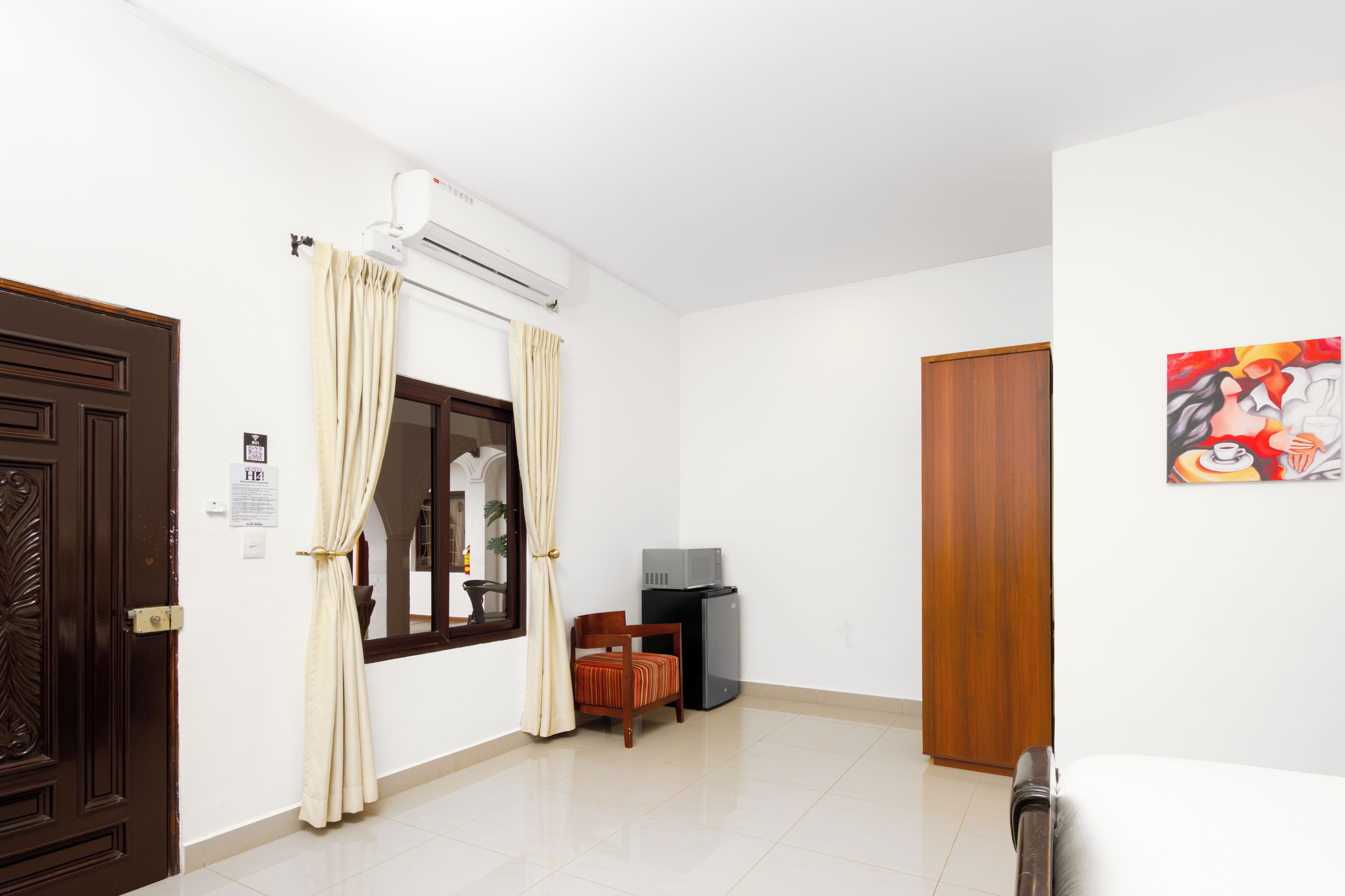 Habitación de hotel con puerta de madera, cortinas color beige, ventana, una mesita con una silla, microondas y un cuadro en la pared.