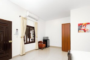 A hotel room with a wooden door, beige curtains, a window, a small table with a chair, a microwave, and a painting on the wall.