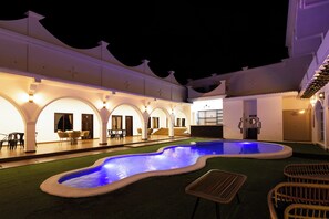 A hotel courtyard with a swimming pool, outdoor seating, and a covered area with stairs leading to a second floor.