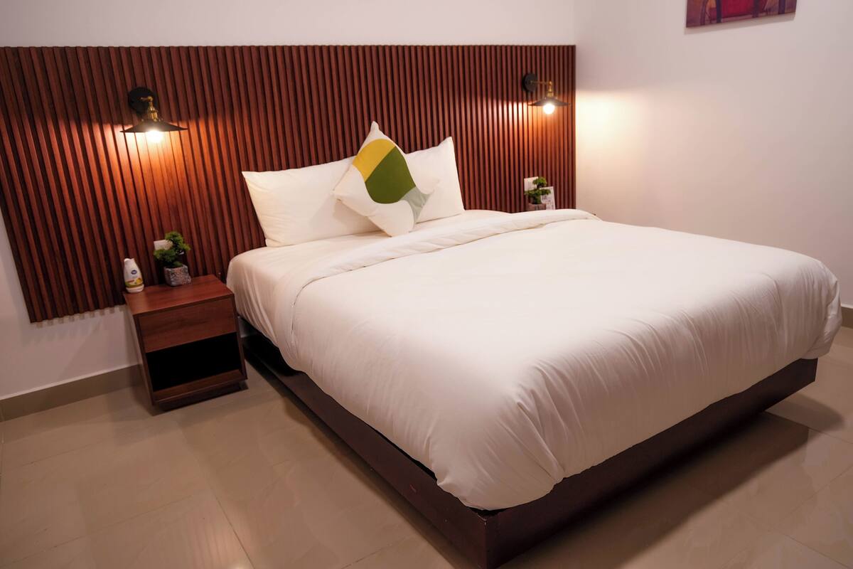 Una cama bien hecha con cabecera de madera, una mesita de noche con una planta y una lámpara de pared.