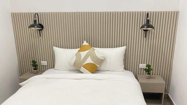 A neatly made bed with a white comforter and a geometric-patterned pillow. There are two bedside tables with lamps, and a small potted plant on each. The room has a striped wall design and a plain white ceiling. - Hotel H4  (El Paraíso)