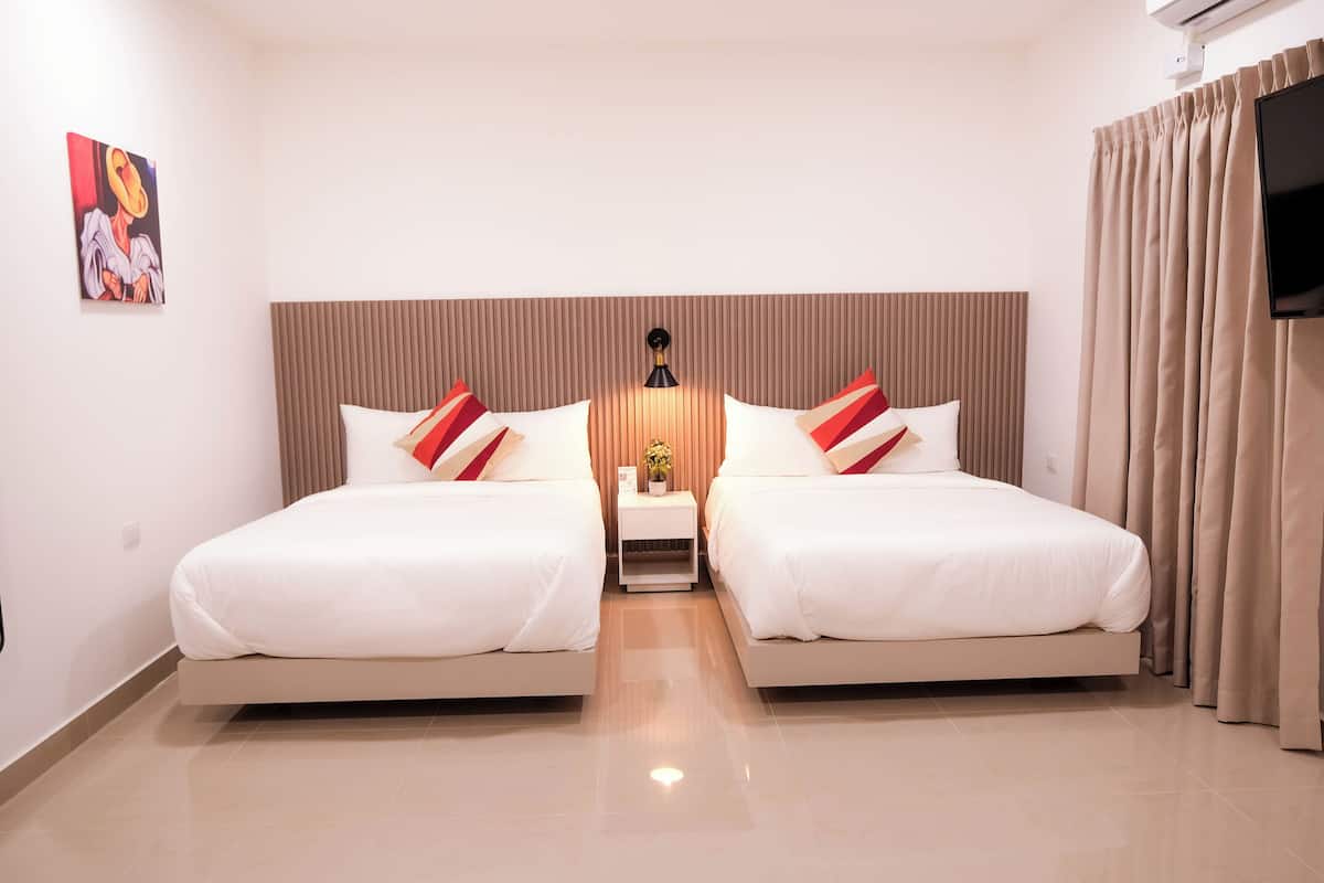 Habitación de hotel con dos camas, un cabecero, una mesita de noche con lámpara, un cuadro en la pared y un televisor a la derecha.