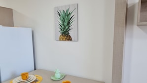 Un coin repas avec une table dressée pour deux personnes, des chaises, une œuvre d’art représentant un ananas accrochée au mur et un tableau blanc.