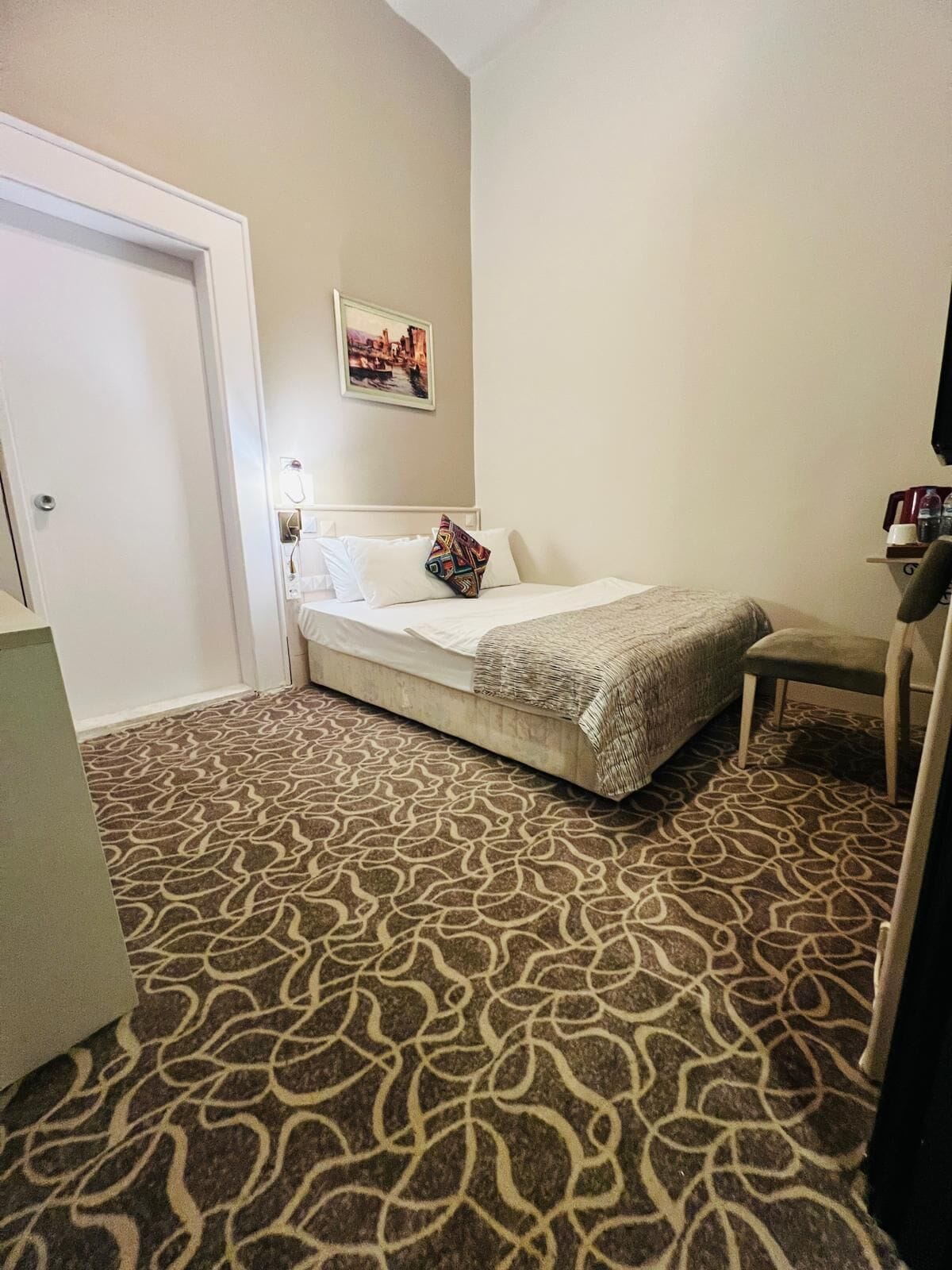 A hotel room with a patterned carpet, a bed with white bedding and a colorful pillow, a small table with a chair, and a wall-mounted TV.