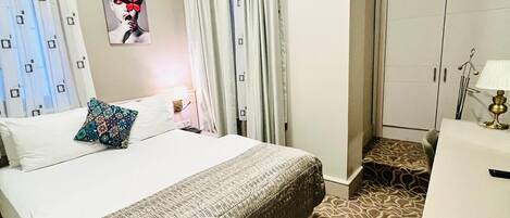A hotel room with a bed, a desk, an air conditioning unit, a wardrobe, and a painting on the wall.