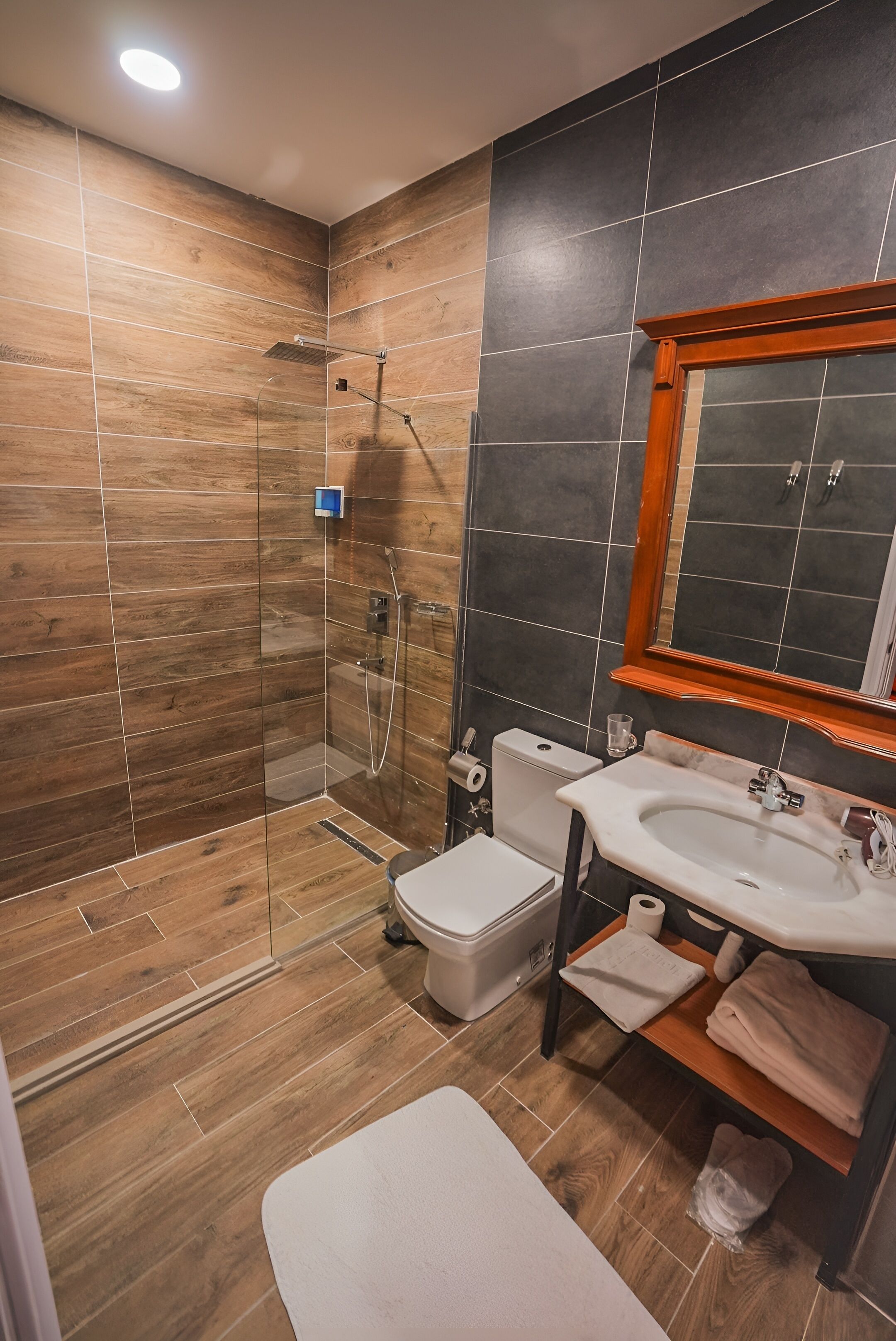 Un baño moderno con ducha de vidrio, inodoro blanco, lavamanos con espejo y suelo de madera.